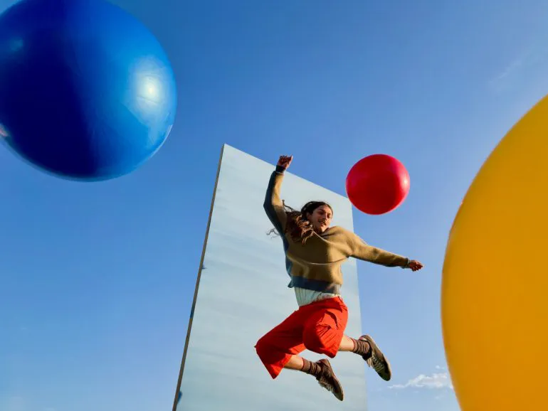 Una persona all’aperto che salta mentre alcuni palloncini colorati volano sullo sfondo di un cielo azzurro e di un fondale grigio rettangolare