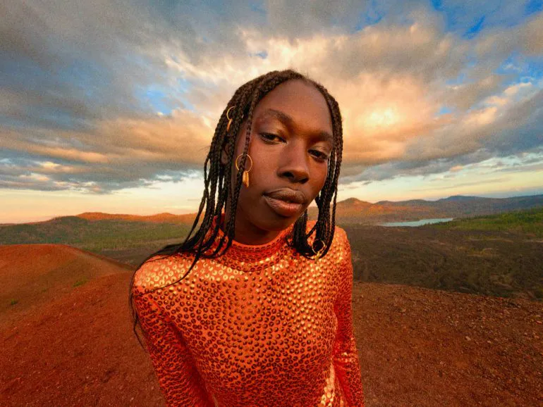 Una persona in posa, vestita di arancione, in un’inquadratura a mezzo busto, sullo sfondo di un cielo nuvoloso e un ampio panorama